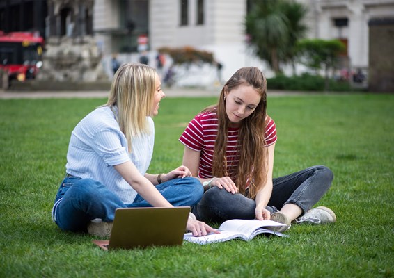 Two female students sitting in the park studying together with a book and a laptop.